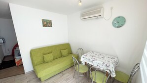 A small, well-lit room with a green sofa, a dining table with chairs, a wall-mounted air conditioner, and a clock. - Apartment Fruk 1 (Jadranovo)