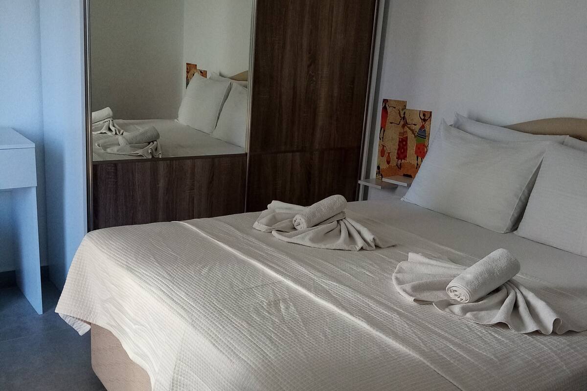 Una cama bien hecha con edredón blanco y dos toallas, cabecera de madera, espejo y un pequeño estante con un adorno.