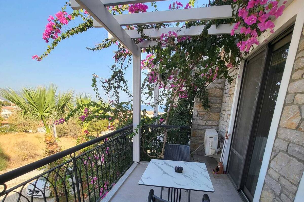 Un balcón con una mesa y sillas blancas, una pérgola con flores rosadas y un muro de piedra.