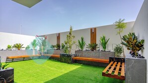 Un patio exterior moderno con césped artificial, bancos de madera y diversas macetas con plantas.