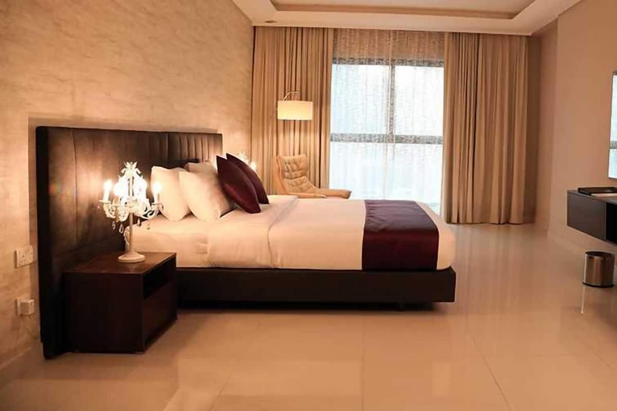 Une chambre d’hôtel moderne avec un grand lit, une table de chevet avec une lampe, une chaise, un meuble pour la télévision et une fenêtre avec des rideaux.