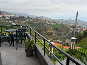 A balcony with a view of a hilly town, a table, chairs, and potted plants.