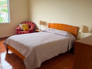 A bedroom with a bed, a chair, two bedside lamps, and a window with a view.