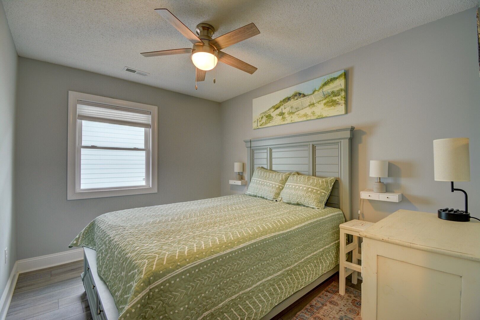 A bedroom with a bed, a ceiling fan, a window, a nightstand, a lamp, and a painting on the wall.