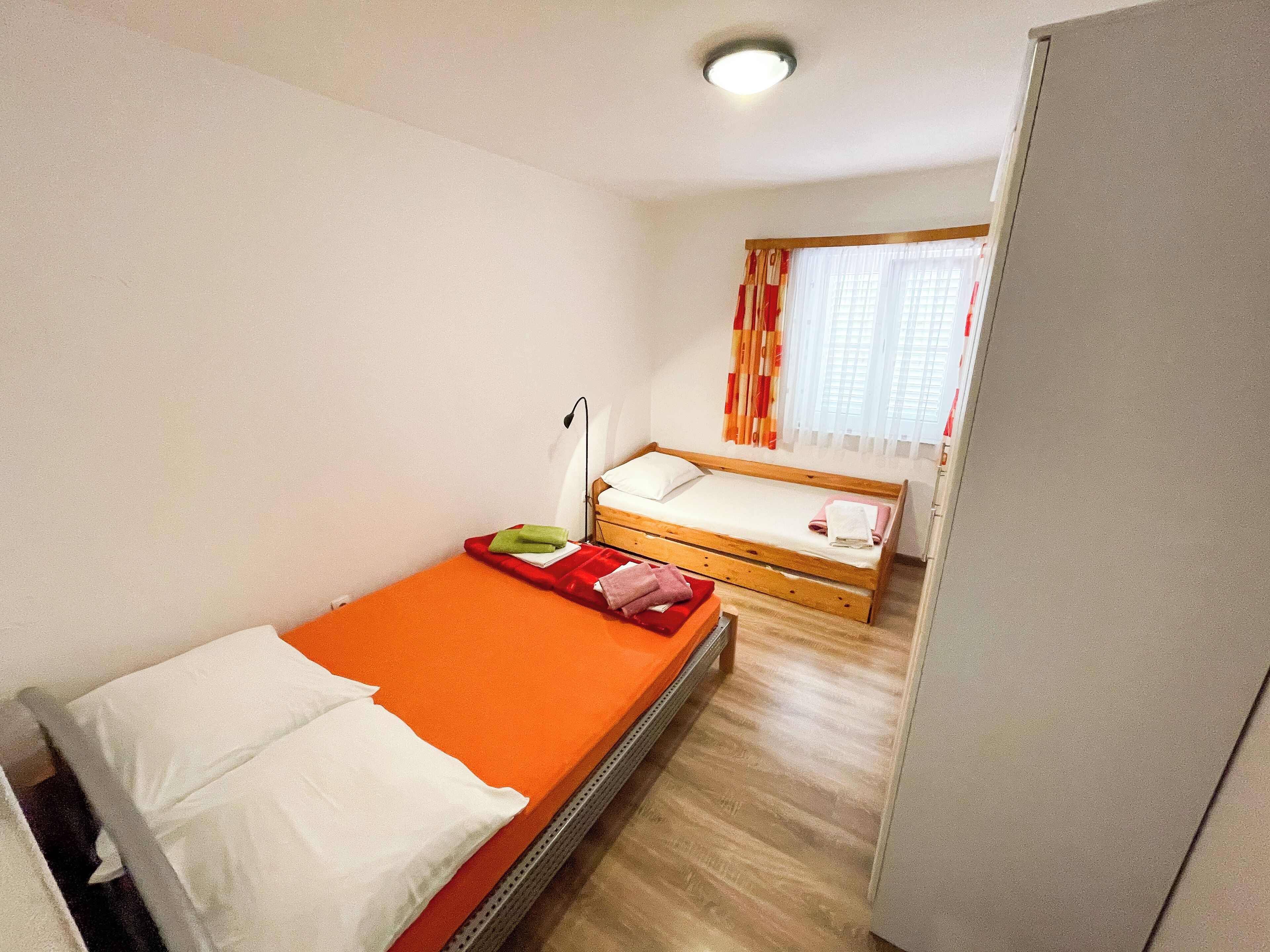 A small, single-bed room with a wooden floor, a bed with an orange mattress, white pillows, and a nightstand with a lamp.