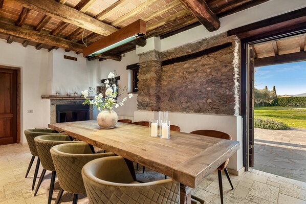 A dining area with a wooden table, cushioned chairs, a vase with flowers, and a stone wall.