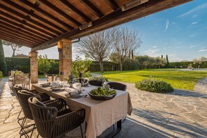 An outdoor dining area with a stone floor, wooden pergola, and a view of a well-manicured lawn and trees.
