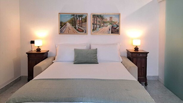 A bedroom with a bed, two bedside tables with lamps, two wall-mounted paintings, and an air conditioning unit. - Casa Romana  (Sanremo)
