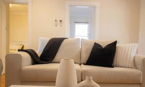 A living room with a beige sofa, two black pillows, a white vase, and a white radiator. - Charming older cottage in Ballarat Central with classic character, modern comfort and a great central location. Two bedrooms, full kitchen, comfy lounge, workspace and easy access to cafés, shops and attractions. (Ballarat Central)