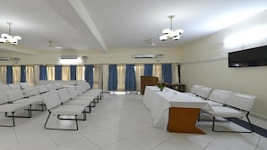 Une salle de conférence avec des rangées de chaises blanches, un pupitre et un téléviseur à écran plat fixé au mur.