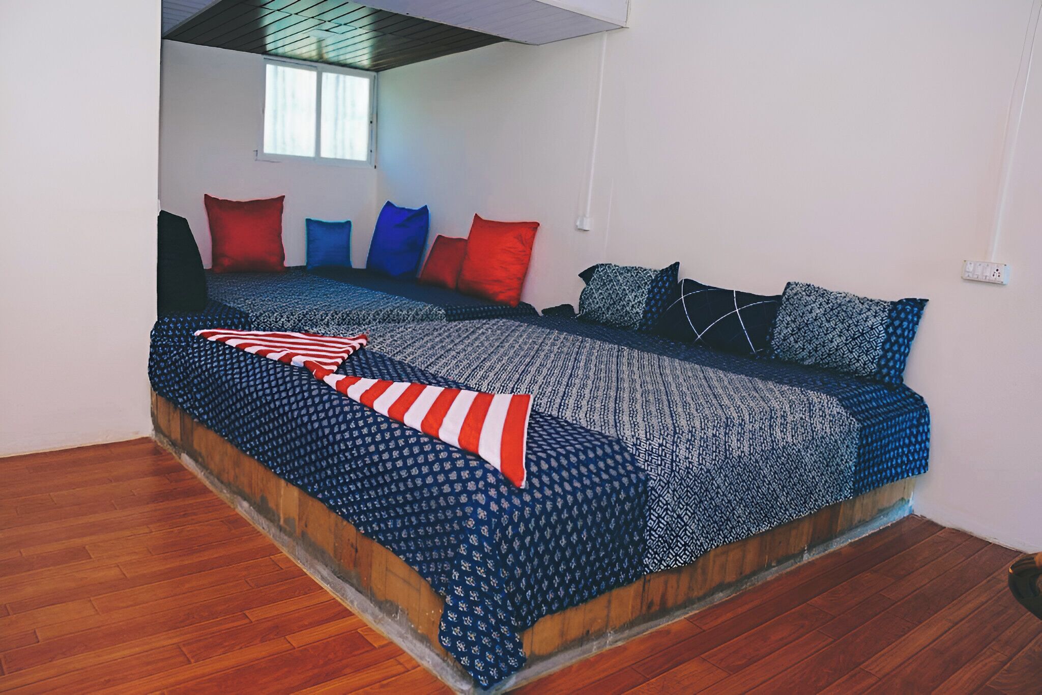A bed with patterned bedding and multiple pillows, a wooden floor, and a window with blinds.