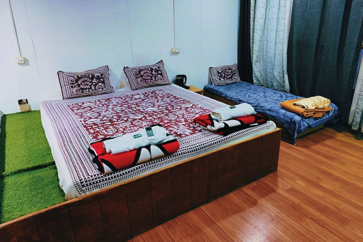 A double bed with patterned bedding, a smaller single bed, a wooden floor, and a green artificial grass area.