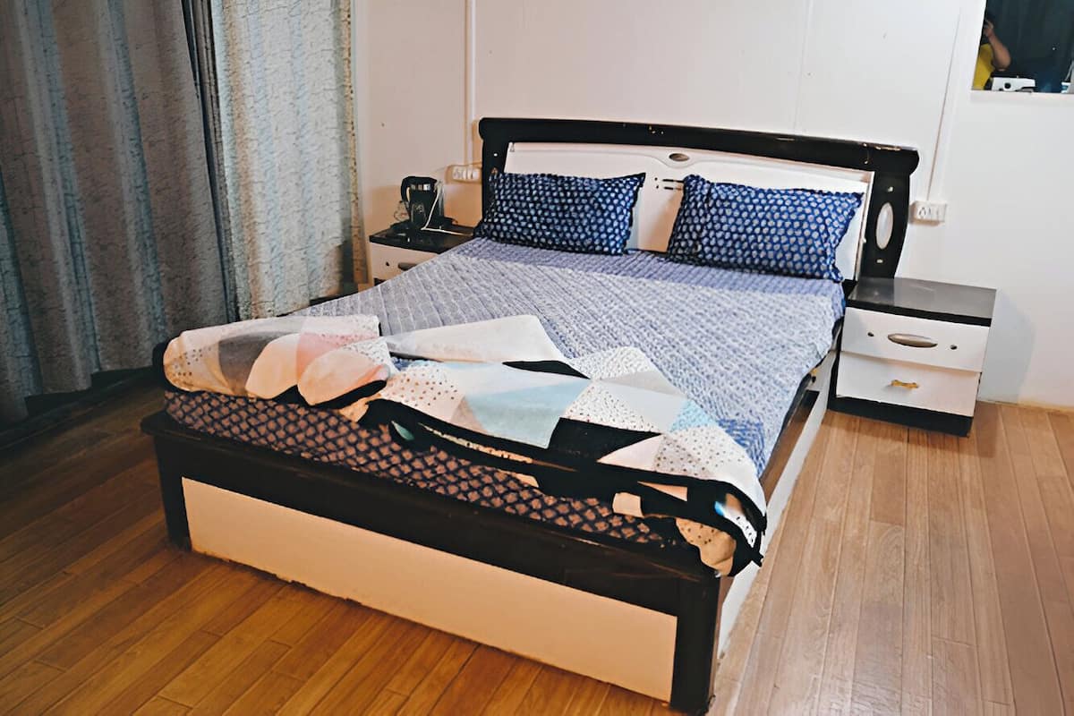 A neatly made bed with a patterned bedspread and pillows, a bedside table with a lamp, and a small drawer unit.