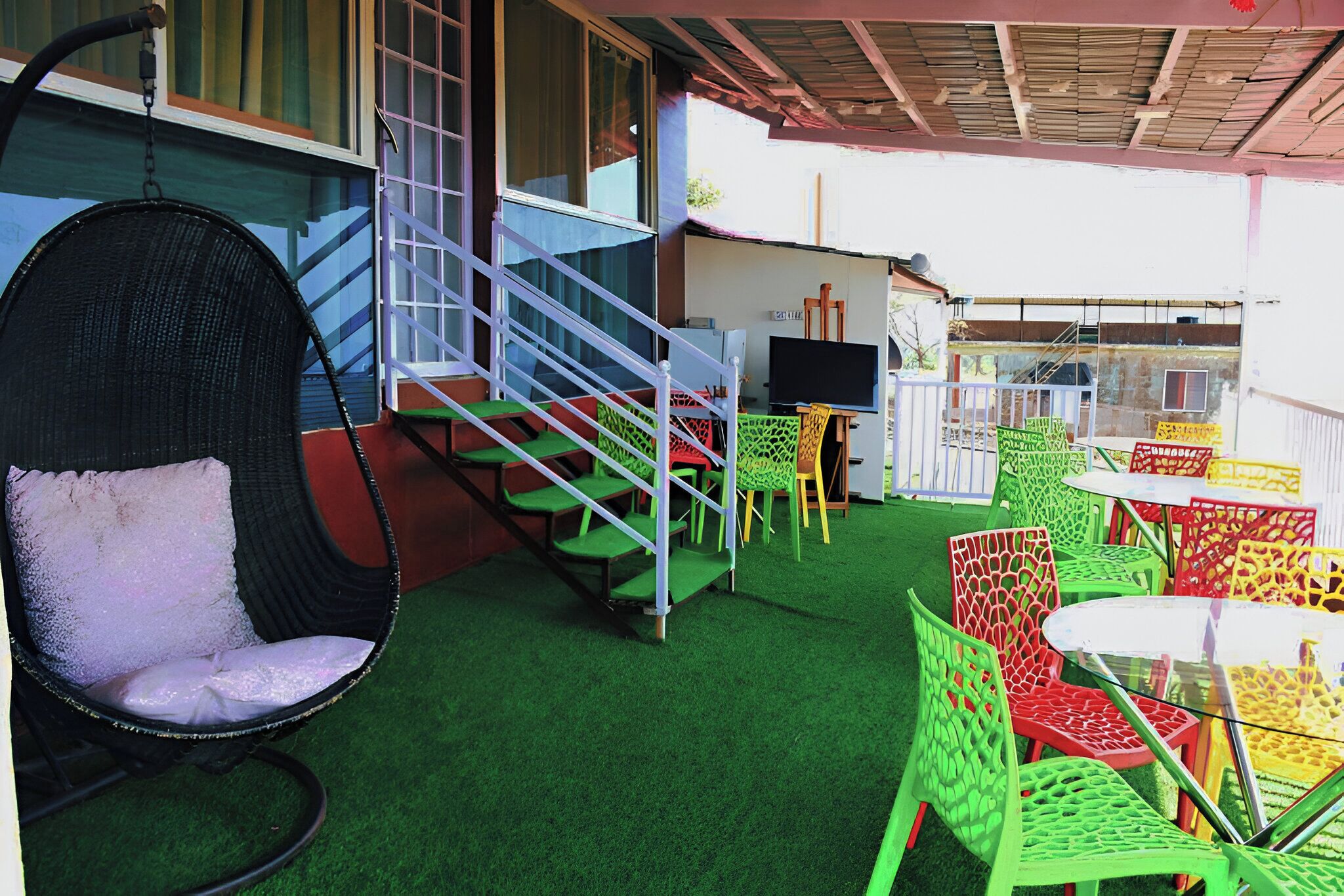A covered outdoor area with a green artificial turf floor, a wicker chair with a cushion, a staircase, and colorful plastic chairs and tables.