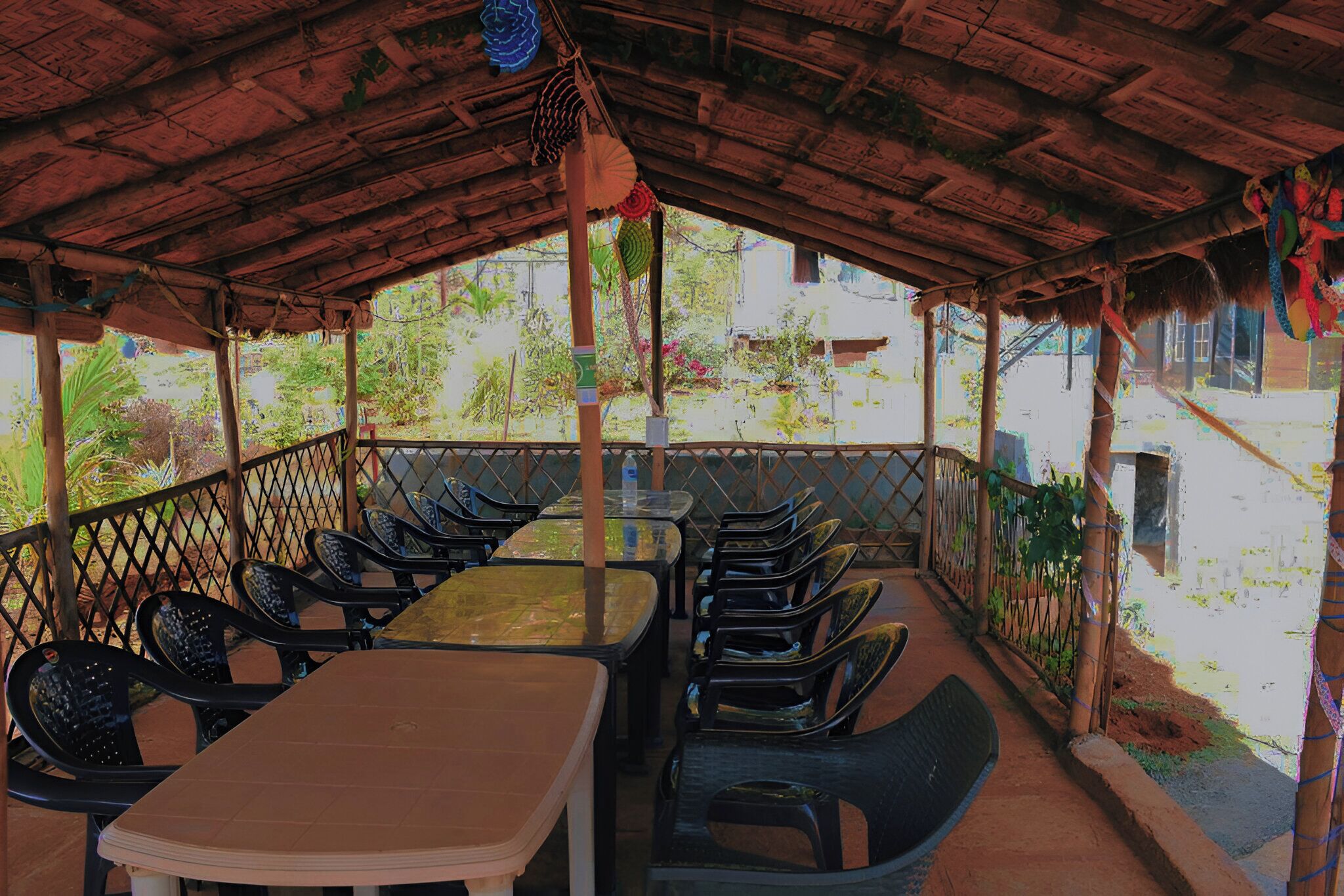 An outdoor seating area with tables and chairs, a thatched roof, and a view of greenery and buildings in the background.