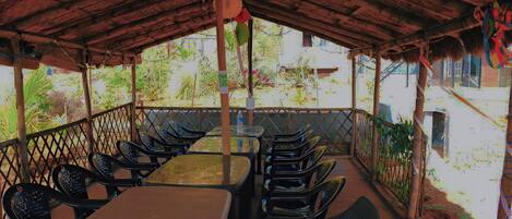 An outdoor seating area with tables and chairs, a thatched roof, and a view of greenery and buildings in the background.