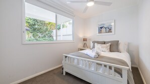 A bedroom with a crib, a large window with a view of greenery, a ceiling fan, and a framed picture on the wall.