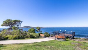 A coastal view with a pathway leading to a deck overlooking the sea, surrounded by greenery and a distant hill.