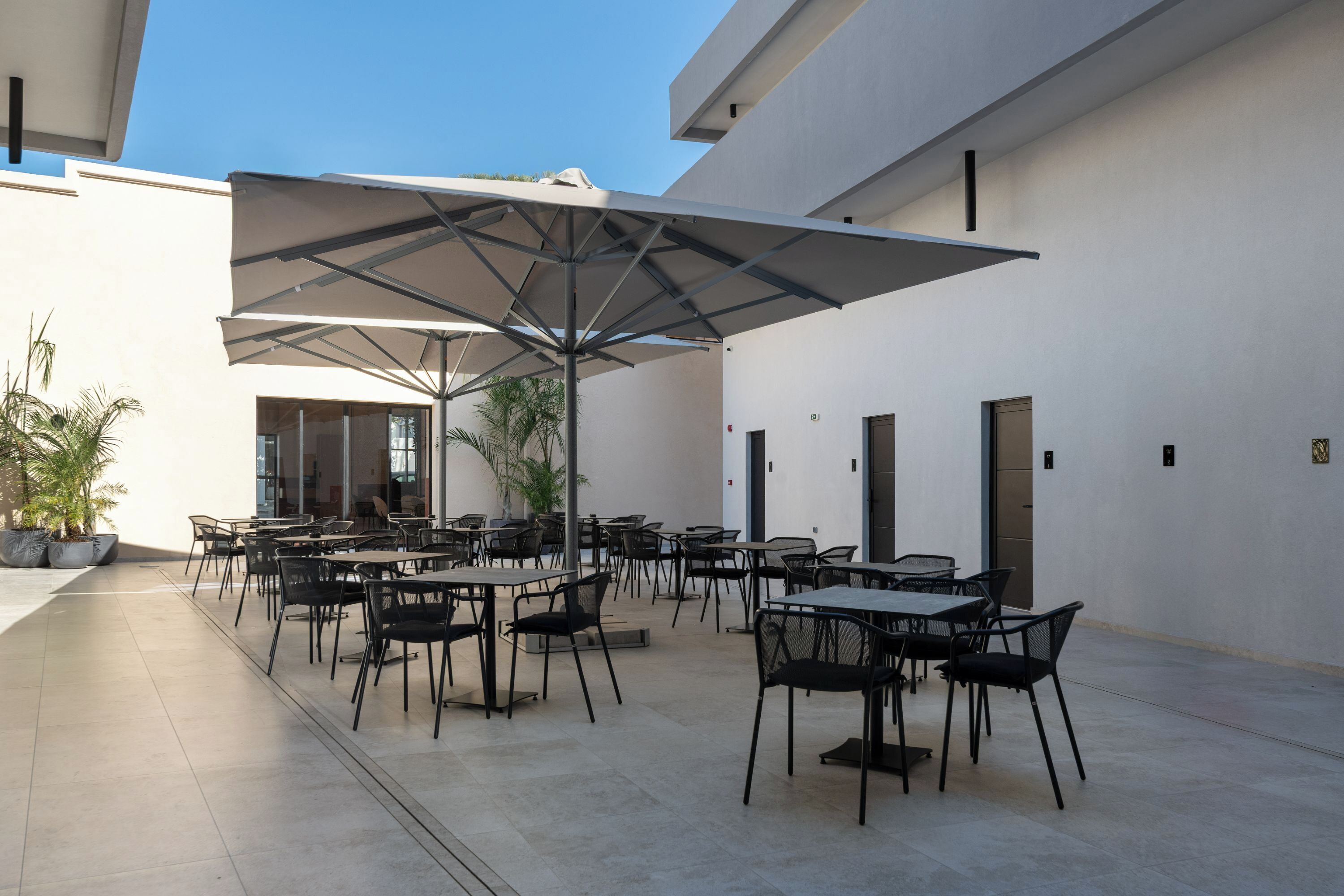 En udendørs patio med flere borde og stole, store parasoller og krukker med planter.