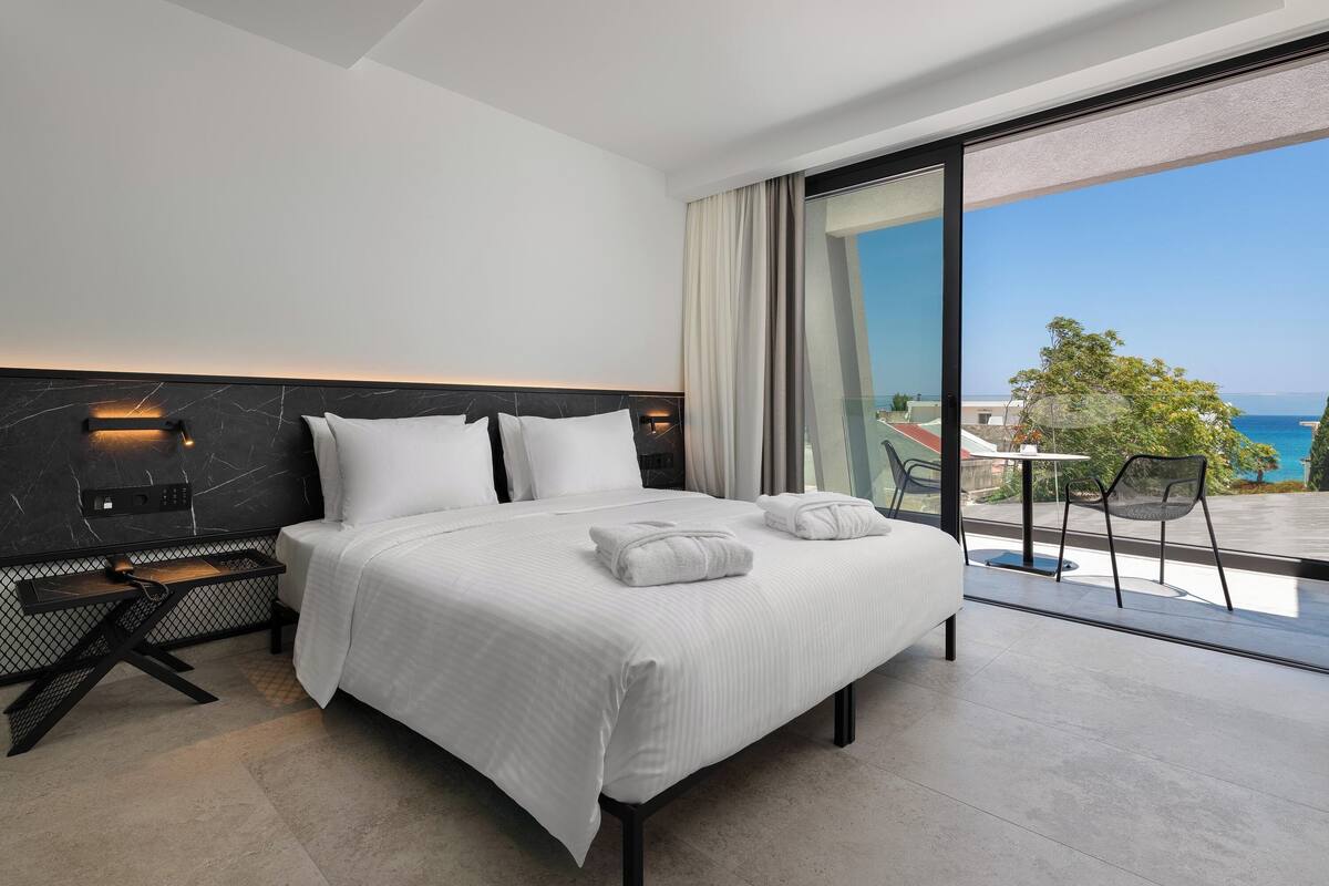 Ein modernes Hotelzimmer mit einem großen Bett, einem kleinen Schreibtisch und einem Balkon mit Meerblick.