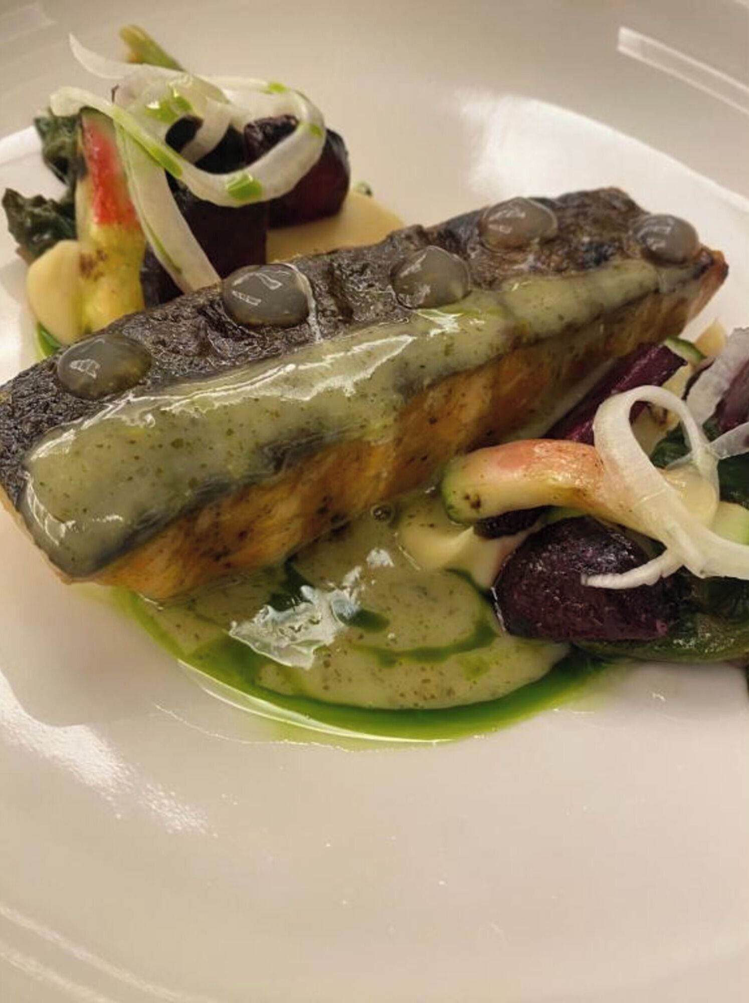 En tallrik med mat innehållande en fiskfilé, oliver och grönsaker.
