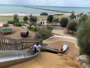 Un parque infantil con tobogán, zona de juegos con arena y vistas a la playa y al muelle.