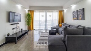 Ein modernes Wohnzimmer mit einer grauen Couch, einem flachbildfernseher an der Wand, einer Topfpflanze und einem großen Fenster mit gelben Vorhängen.