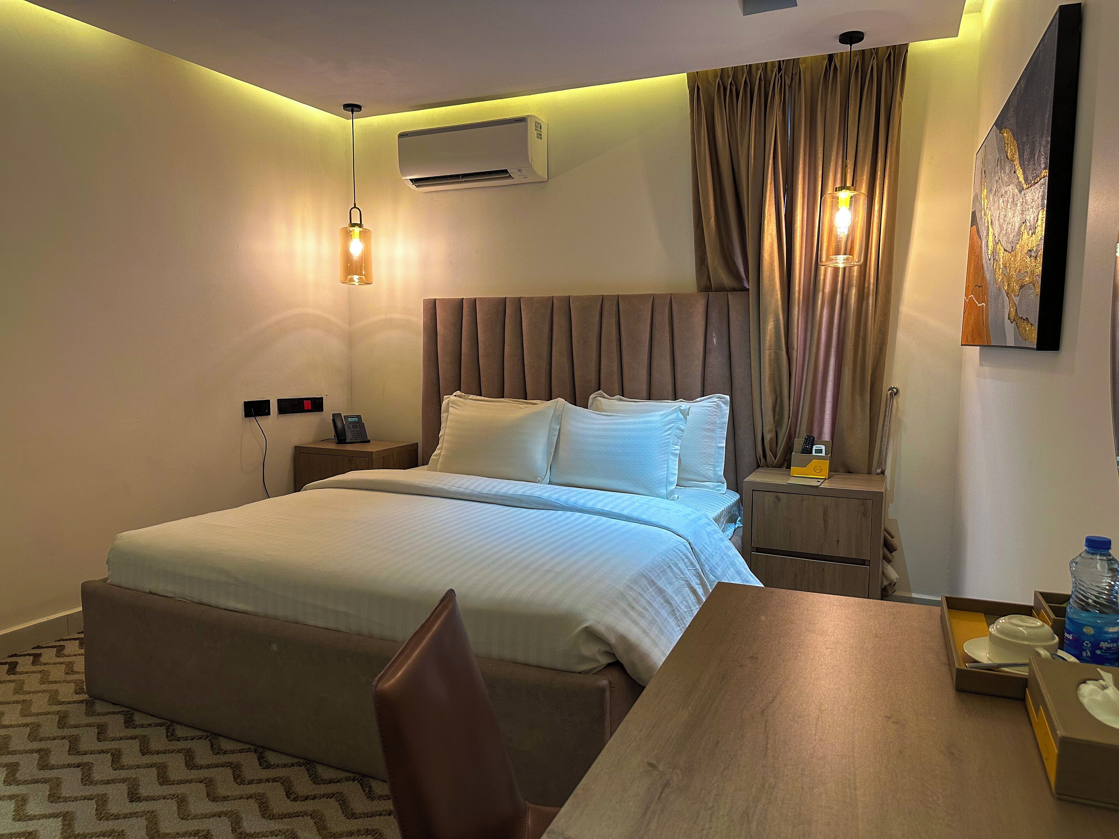 A hotel room with a bed, bedside tables, a desk, a chair, an air conditioning unit, and a painting on the wall.