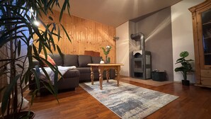 A cozy living room with a wooden wall, a sofa, a table, and a wood-burning stove. - Cozy vacation apartment in an idyllic natural location (Rosenthal-Bielatal)