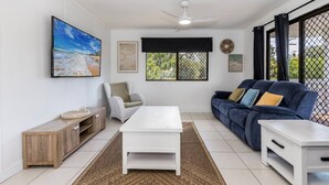 A living room with a white coffee table, a blue sofa, a wicker chair, a wooden cabinet, and a large framed picture on the wall. - A Lure - 100m to Main Beach! (Agnes Water)