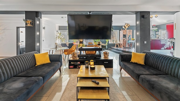 Una sala moderna con un televisor de pantalla plana grande, dos sofás con cojines amarillos, una mesa de centro y un área de comedor al fondo.