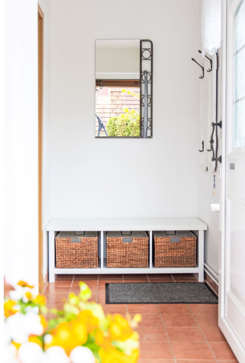 A clean, modern entryway with a white bench, three wicker baskets, a mirror, and a door with a metal handle.