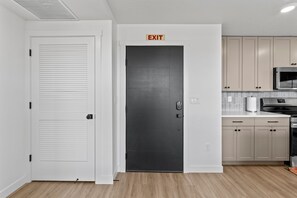 Una cocina moderna con suelo de madera clara, armarios blancos, un microondas de acero inoxidable y una señal de salida sobre una puerta oscura.