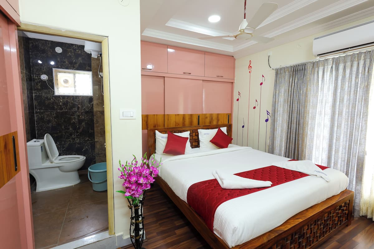 A hotel room with a bed, a wooden headboard, a red and white bedspread, a vase with flowers, a ceiling fan, and a bathroom with a toilet and sink.