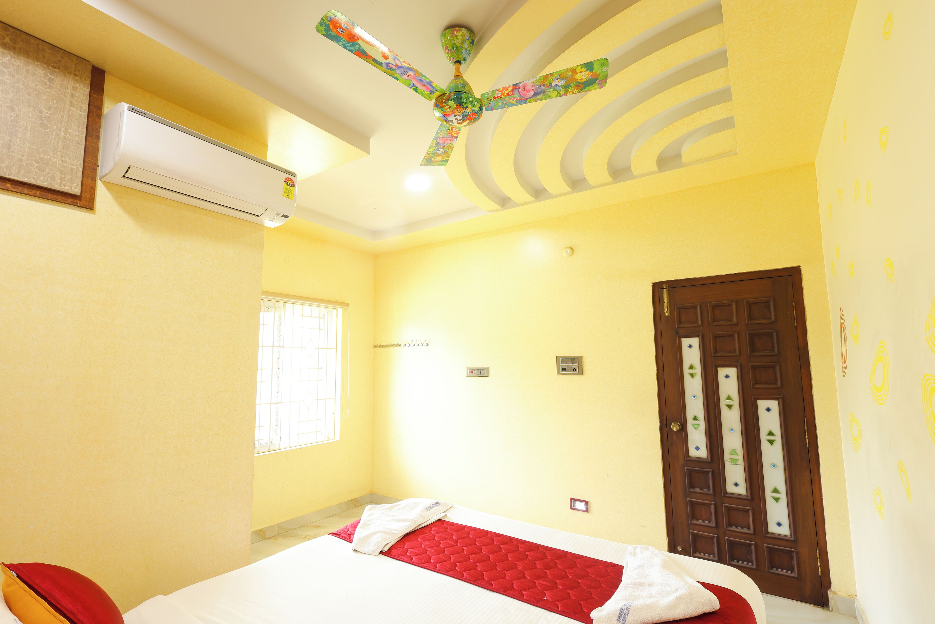A room with a bed, an air conditioner, a decorative ceiling fan, a window, and a door with decorative glass panels.