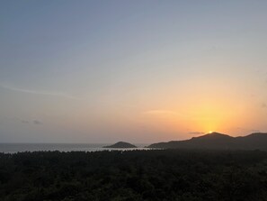 A sunset over a coastal landscape with a forested hill in the foreground. - The Nautical Nest (Canacona)
