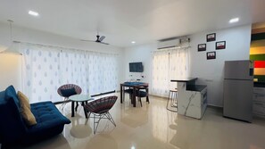 A modern living room with a blue sofa, a dining table with chairs, a TV mounted on the wall, and a refrigerator. - The Nautical Nest (Canacona)
