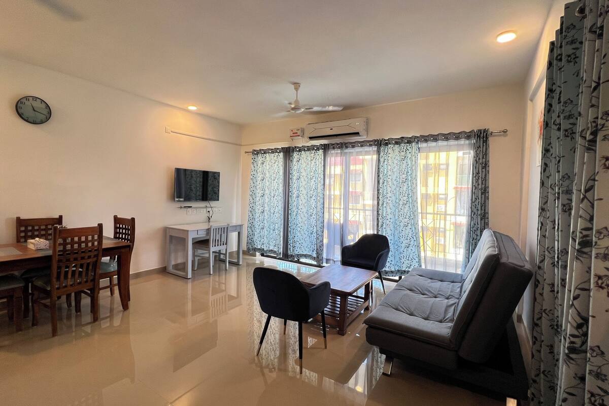 A living room with a sofa, a dining table with chairs, a TV mounted on the wall, and a ceiling fan.