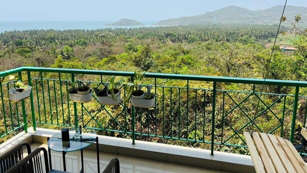 A balcony with a glass table and chairs, potted plants, and a view of a lush landscape with mountains and a body of water. - The Nautical Nest (Canacona)