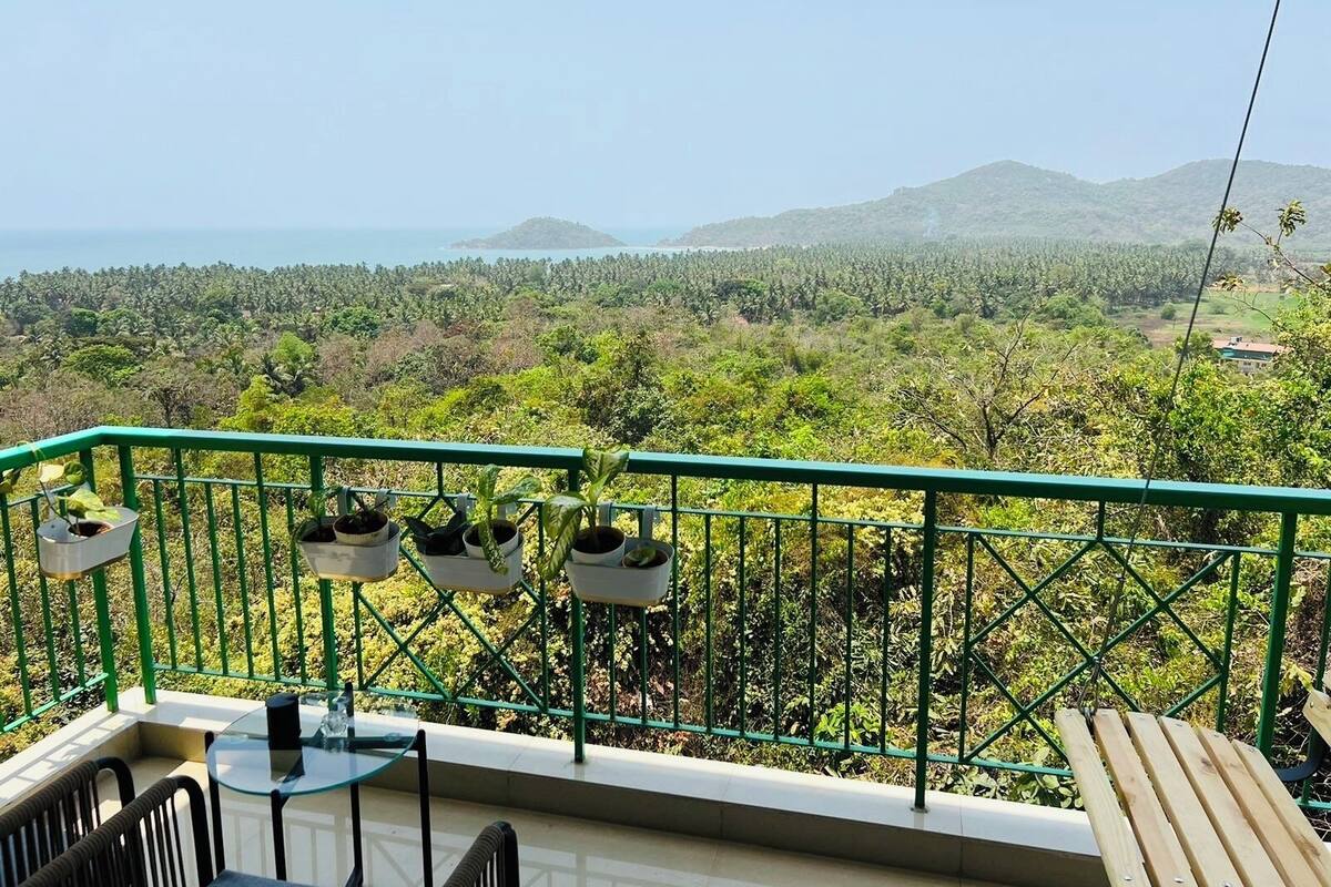 A balcony with a glass table and chairs, potted plants, and a view of a lush landscape with mountains and a body of water.