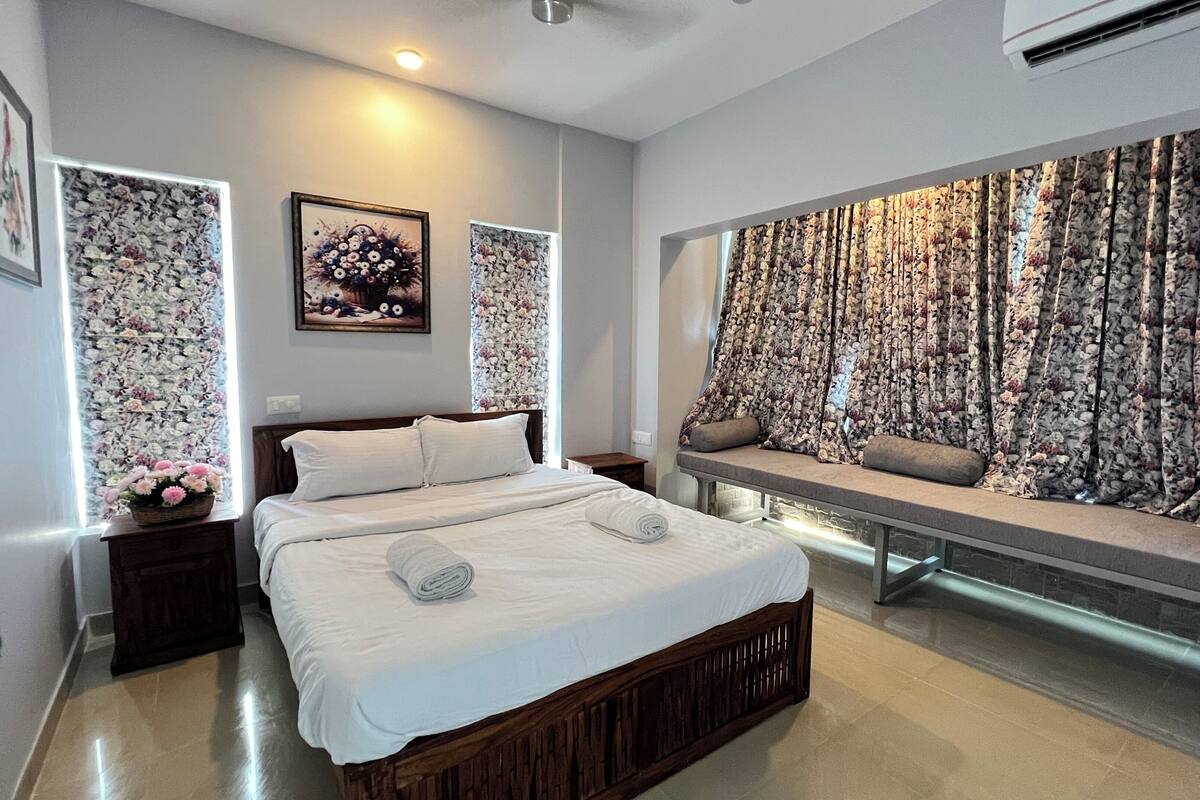 A bedroom with a large bed, a bench with cushions, floral curtains, and framed artwork on the walls.