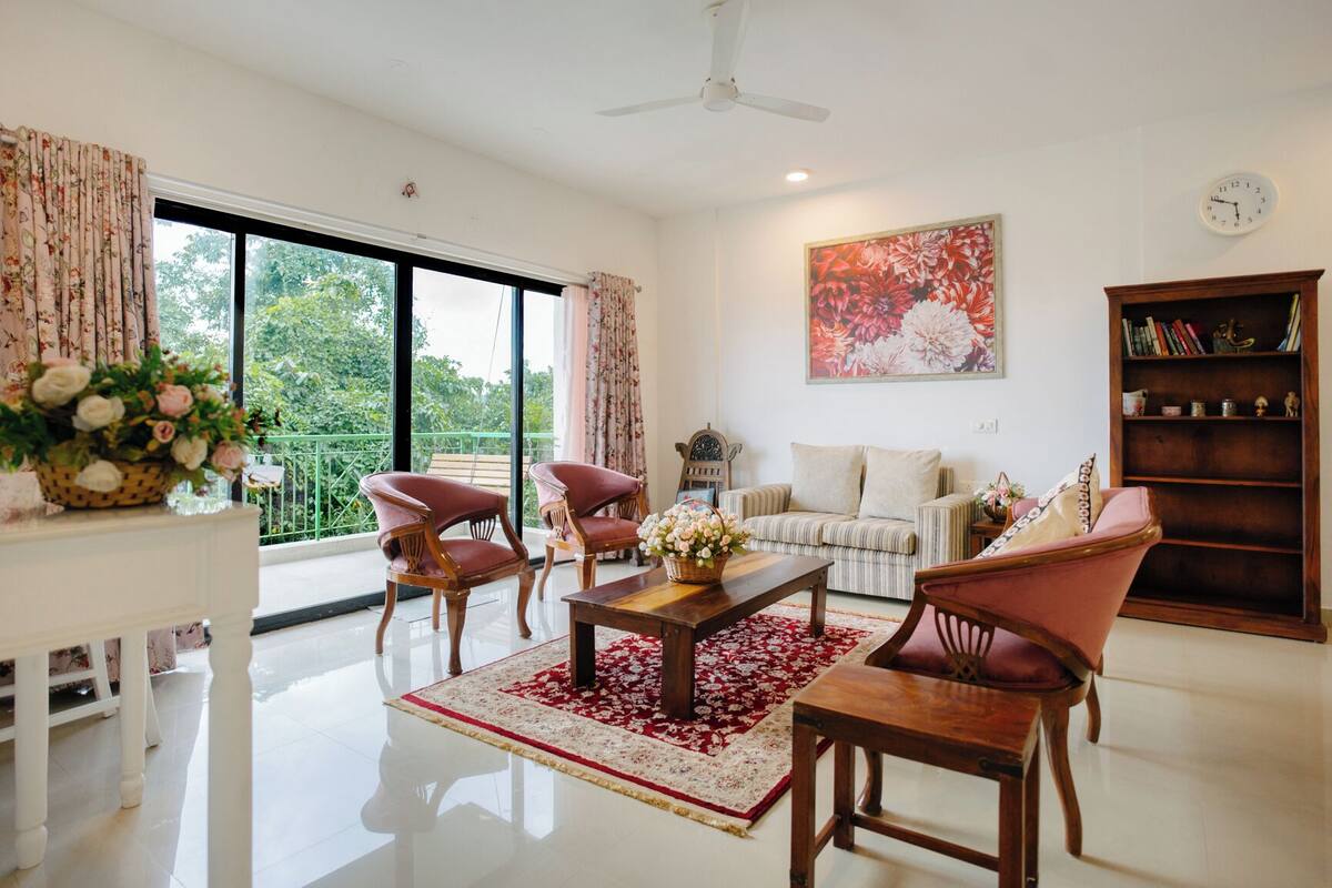 A spacious living room with a large window, a ceiling fan, a bookshelf, and a floral painting.