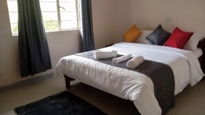 A neatly made bed with white linens and a dark-colored blanket, a black rug on the floor, and a window with curtains. - MT LONGONOT COUNTRY RESORT NAIVASHA (Naivasha)