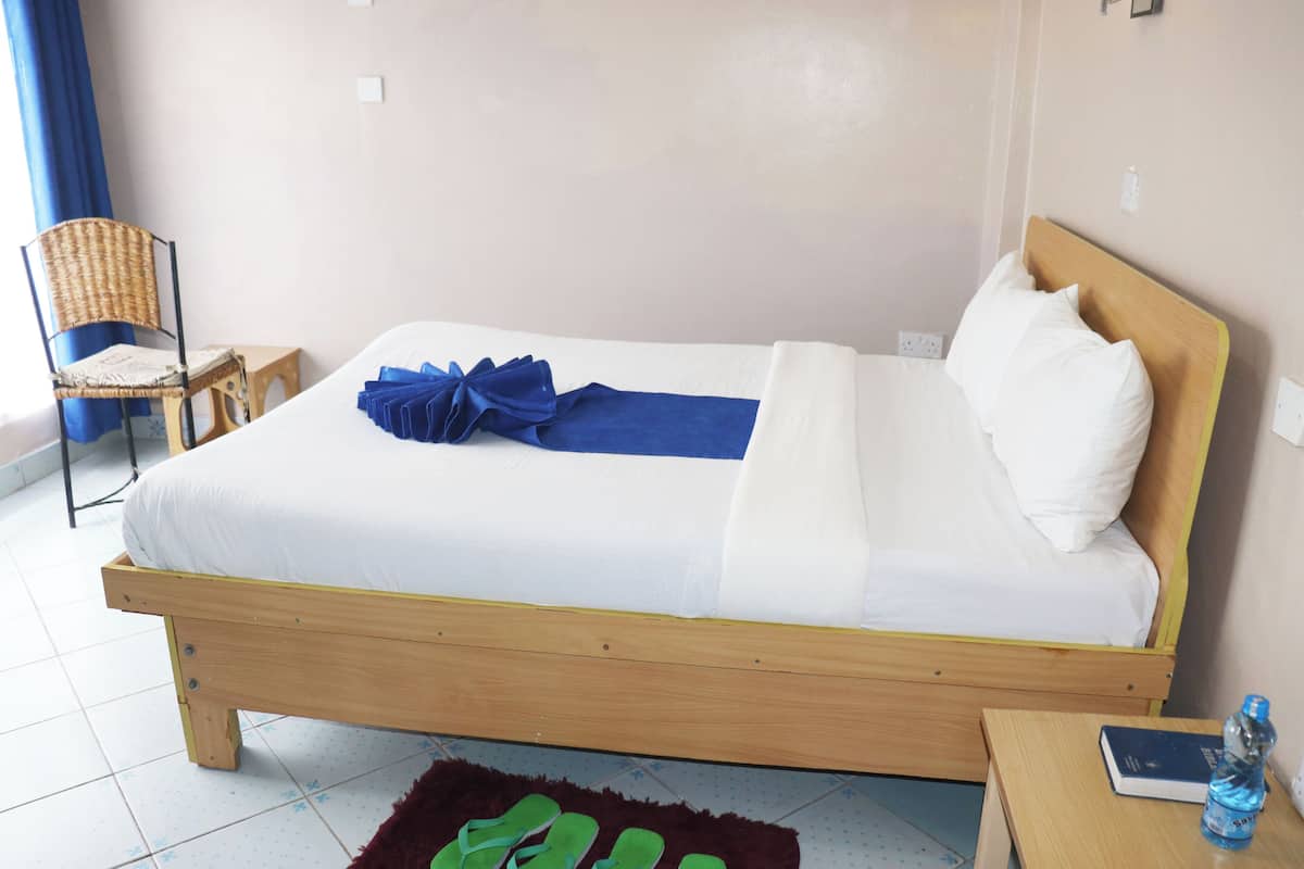 A single bed with a wooden frame, white bedding, a blue towel, and a wooden nightstand with a book and a water bottle.
