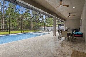 A covered outdoor swimming pool area with a view of trees, a patio with chairs and a table, and ceiling fans.