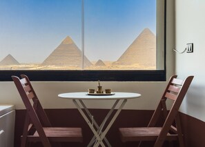 A room with a window view of the pyramids, a small round table with a tray, and two wooden chairs. - Xuru Stays I Jacuzzi Studios @ The Pyramids (Giza)