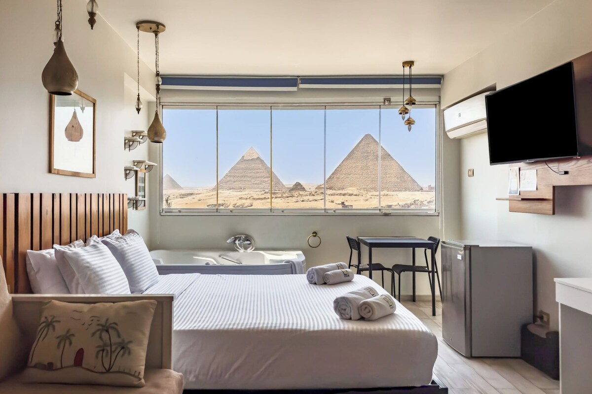 A hotel room with a bed, a desk, a chair, a TV, and a view of the pyramids.