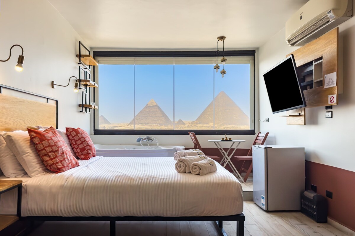 A hotel room with a large bed, a small table, a TV, and a view of pyramids.