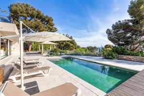 Piscina com espreguiçadeiras e guarda-sóis, rodeada por árvores e um muro de pedra.