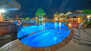 Une piscine nocturne dotée d’une terrasse en bois, de palmiers et de structures à toits de chaume.